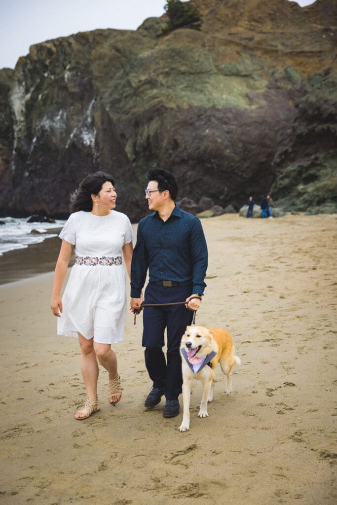 Couple with their dog enjoying walk on the beach during a photoshoot, captured in a natural and candid moment by a San Francisco Bay Area wedding photographer.